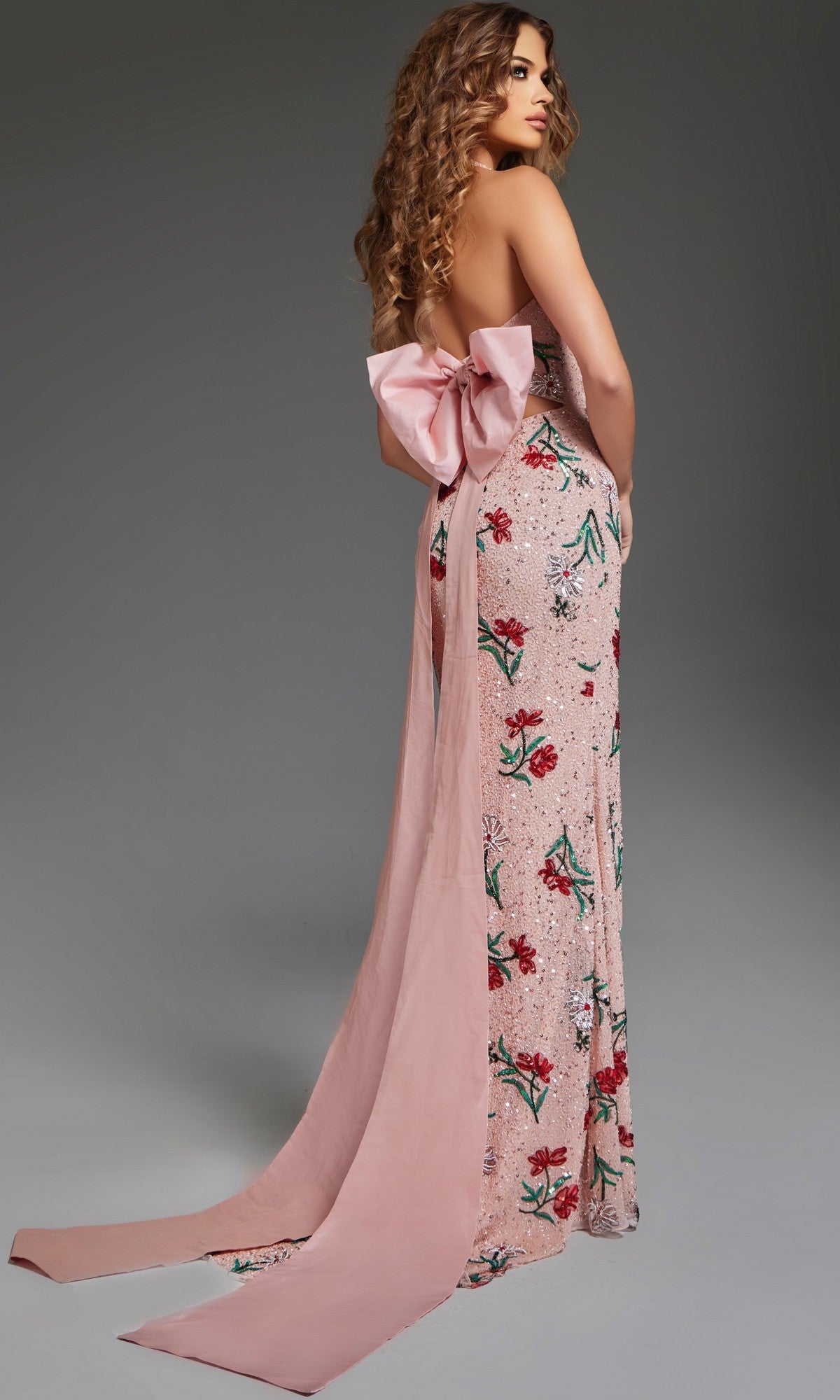 A woman stands against a plain gray background wearing the Jovani 44636 long prom dress, featuring a floral print, strapless design, and a large pink bow at the back.